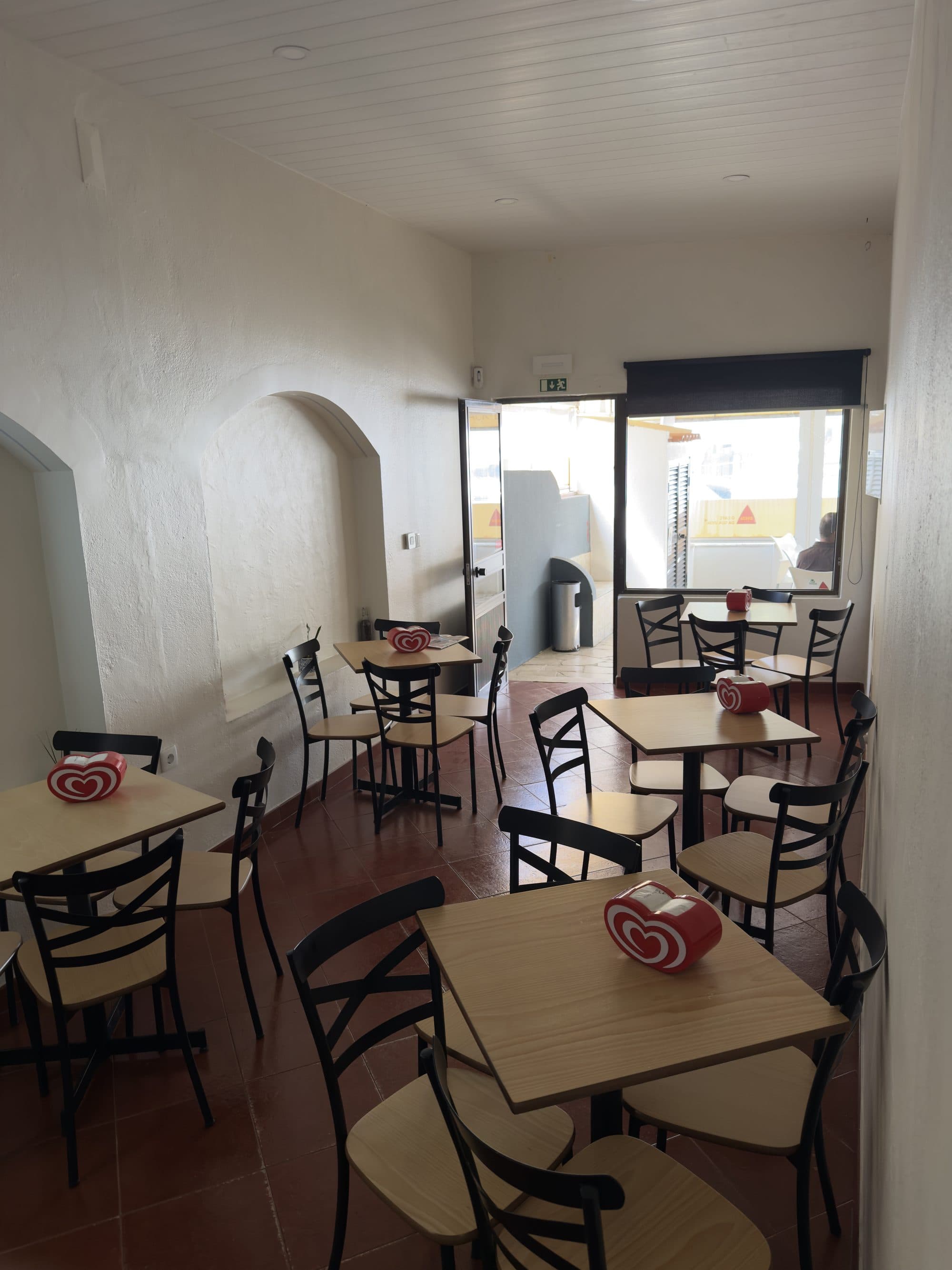 Sala de refeições clara, com mesas de madeira e porta aberta para a rua